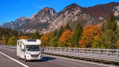 An RV travels along a scenic highway, accentuated with mountains and green trees in the background.