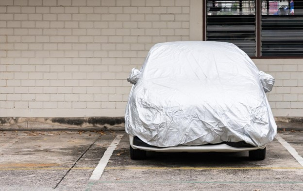 A white sedan sits in a parking lot under a vehicle cover, shielded from the elements.