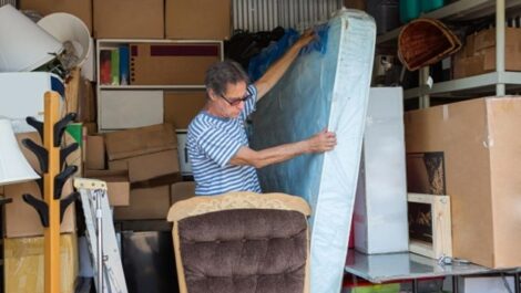 A man pulls his mattress out of a full storage unit, stacked high with boxes. His mattress is wrapped in a protective plastic cover.