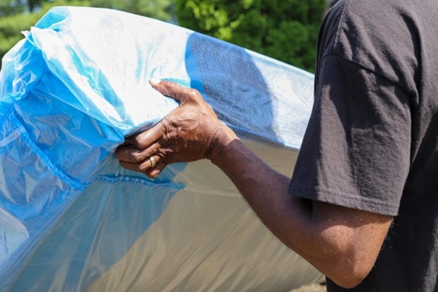A man lifts a mattress from its short end as he begins to transport it. The mattress is inside of a fabric mattress cover.