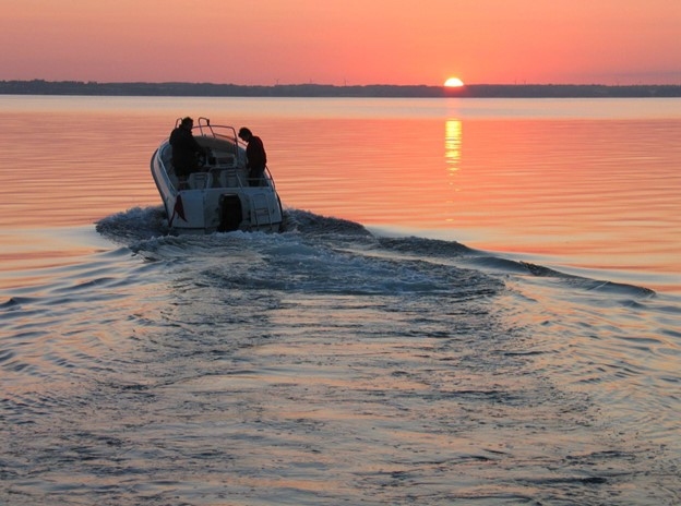 A white speed boat cruises on the waters of a lake, with a red, yellow, and orange sunset in the background.