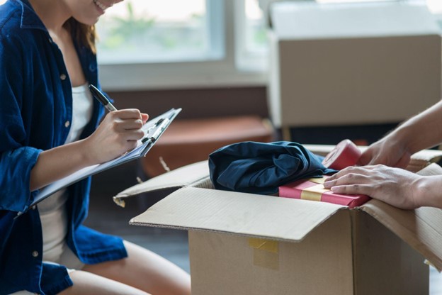 A woman writes on a checklist while their partner packs a box full of belongings.