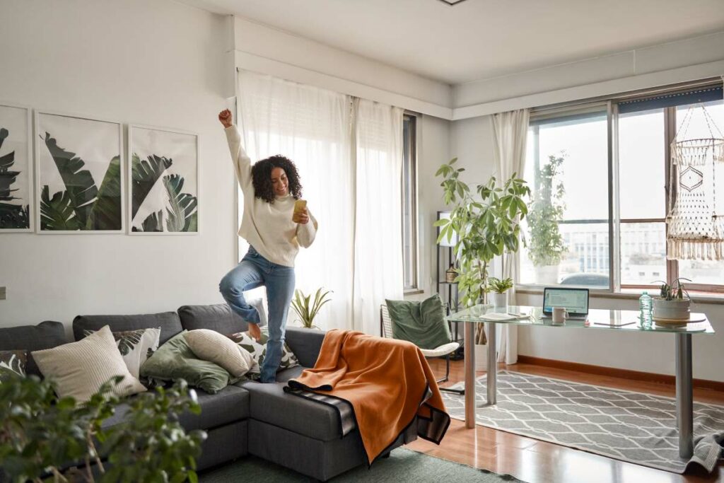 Happy young woman listening to music dancing in her clean, decluttered apartment.